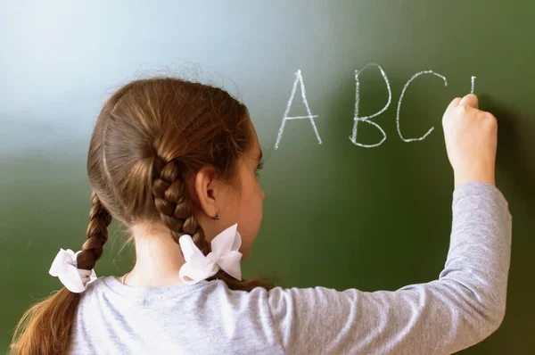 Basisschool meisje staat voor een bord en schrijft met krijt — Stockfoto