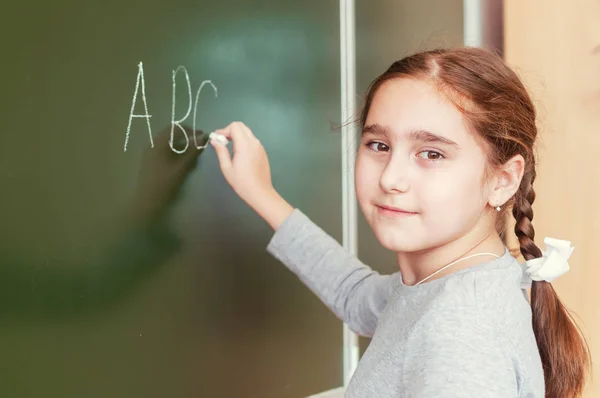 Basisschool meisje staat voor een bord en schrijft met krijt — Stockfoto
