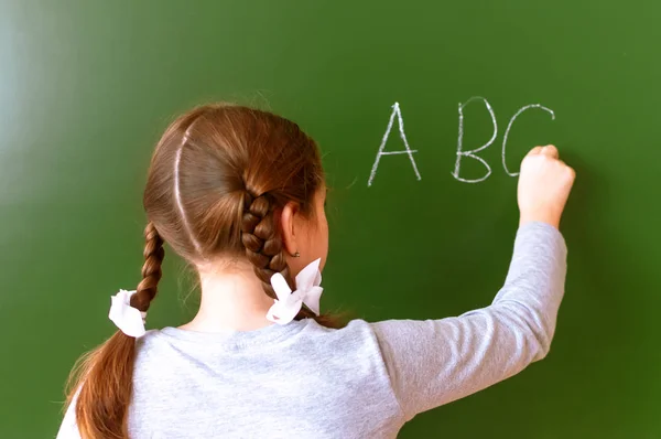 Basisschool meisje staat voor een bord en schrijft met krijt — Stockfoto