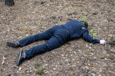 Ormanda cinayet. Mavi gömlekli ve pantolonlu bir adamın cesedi ormandaki ağaçların arasında yerde yatıyor. Bir saldırı kurbanı. Yatay fotoğraf.