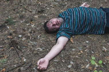 Ormanda cinayet. Mavi tişörtlü bir adamın cesedi ormandaki ağaçların arasında yerde yatıyor. Bir saldırı kurbanı. Yatay fotoğraf.