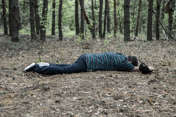 Murder in the woods. The body of a man in a blue t-shirt and trousers lies on the ground among the trees in the forest. Victim of an attack. Horizontal photo.