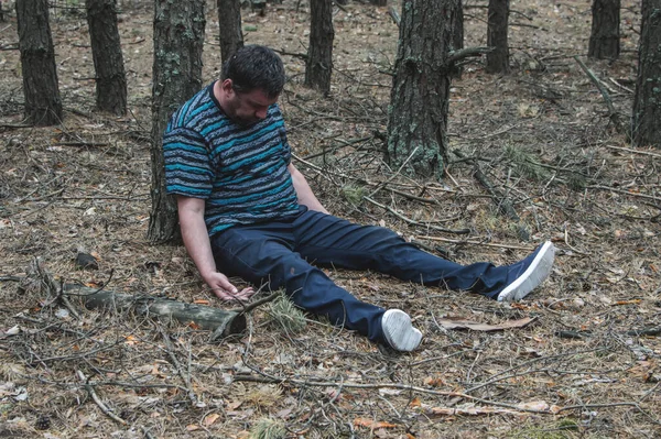 Murder in the woods. The body of a man in a blue t-shirt and trousers is sitting on the ground among the trees in the forest. Victim of an attack. Horizontal photo. Close up.