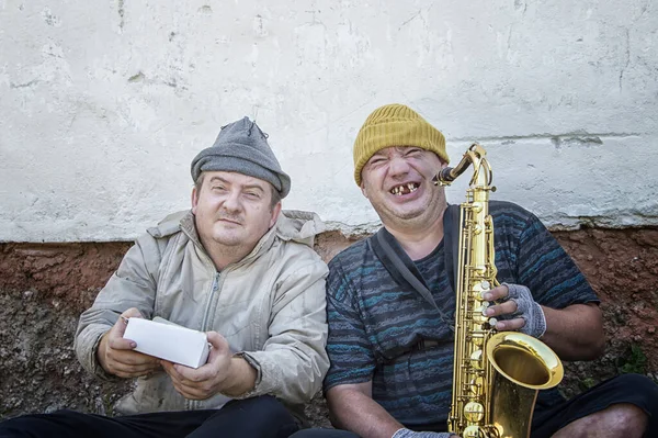 Homeless street musicians sit on the street with a saxophone and a donation box. Poverty and hunger.