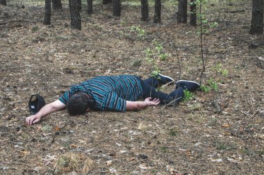 Ormanda cinayet. Mavi tişörtlü ve pantolonlu bir adamın cesedi ormandaki ağaçların arasında yerde yatıyor. Bir saldırı kurbanı. Yatay fotoğraf.