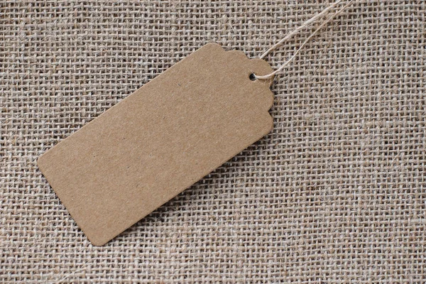 Empty paper tag with rope on the background of coarse fabric. Recycled cardboard tag. Price tag for discounts, gift tag, sale tag, tag for agricultural products