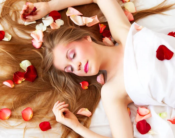 Красивая сексуальная девушка с длинными волосами расслабляется на белых простынях с лепестками роз — стоковое фото