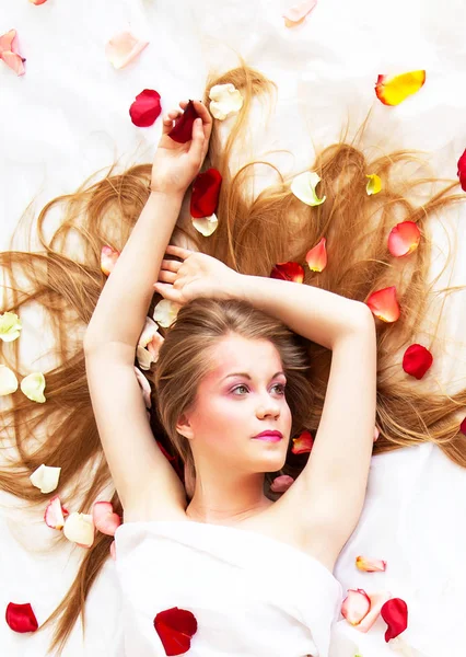 Beautyful сексуальна дівчина з довгим волоссям, що розслабляється на білих простирадлах з пелюстками троянд — стокове фото