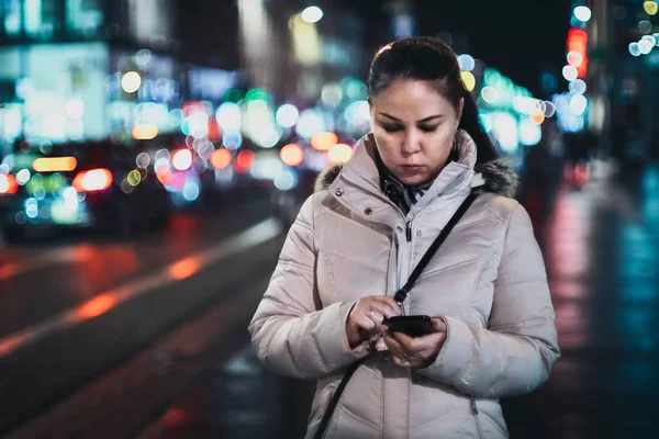 A girl looks at a mobile phone on a street of a big city at night, against the background of the lights of passing cars and street lighting