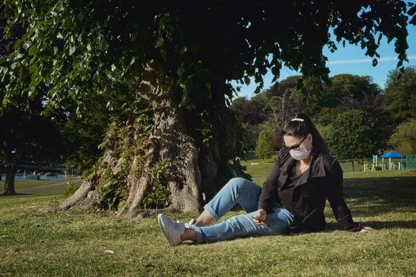 A woman in a protective mask is sitting on grass in the park