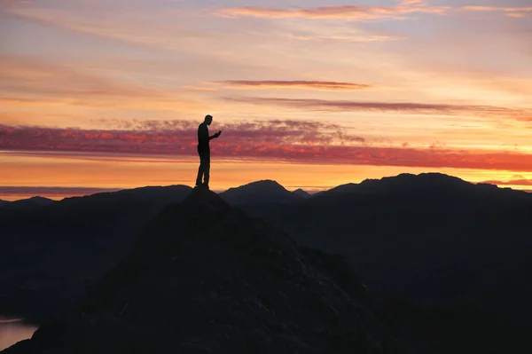 Dağın tepesinde akıllı telefon kullanan bir adam silueti çarpıcı bir günbatımı gökyüzünün arka planına karşı. Lomond Gölü ve Trossachs Ulusal Parkı, İskoçya