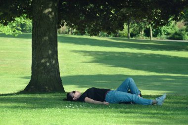 Parktaki çimlerin üzerinde bir kadın yatıyor. Büyük ağacın yanında.