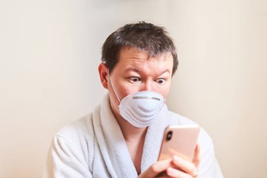 Tıbbi maskeli ve sabahlıklı bir adam telefona şaşkın gözlerle bakar, kendini izole eder, karantina yapar, evde kalır, online sipariş verir.