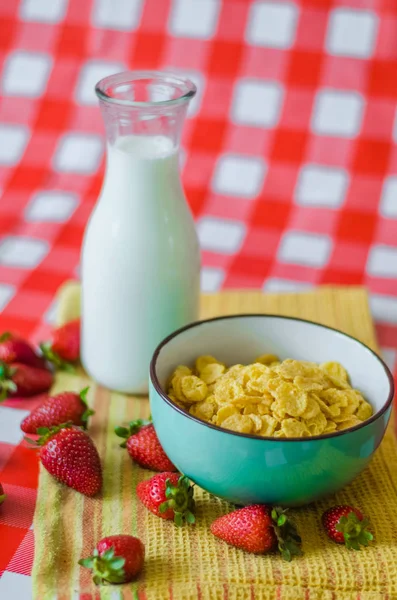 Fresh milk in glass bottle, cereals in green blue ceramic bowl, tasty yogurt in small glass bowl with a lot of strawberries around, on yellow kitchen towel