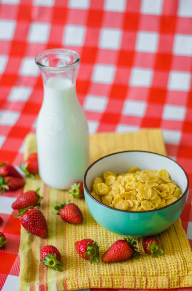 Fresh milk in glass bottle, cereals in green blue ceramic bowl, tasty yogurt in small glass bowl with a lot of strawberries around, on yellow kitchen towel