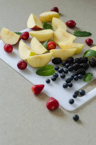 Sliced fresh apples, strawberries, blueberries and blackberries, mint leaves around, fresh summer fruits