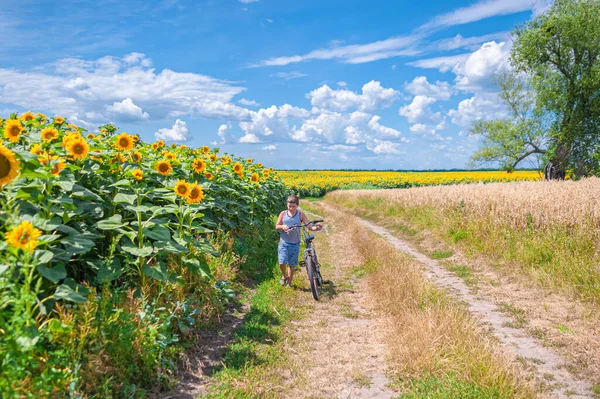 A young boy in a T-shirt and shorts with a bicycle walks along a rural road between a field of sunflowers and trees in the forest