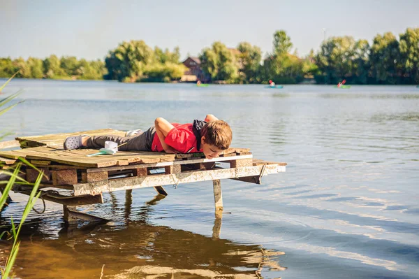 stock image a boy lies on a fishing bridge with a toy boat and looks into the water of the lake