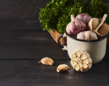 Taze Çiğ sarımsak ampuller ve karanfil beyaz Kupası ve siyah ahşap zemin üzerine yeşil karışık yaprak