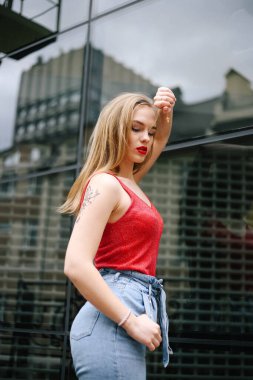 Cam bir binanın arka planında kırmızı tişört ve kot pantolon giymiş güzel bir kız model. Sokak tarzı, moda, yaşam tarzı