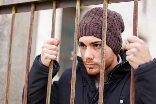 attractive young man in coat and knitted cap posing behind the bars