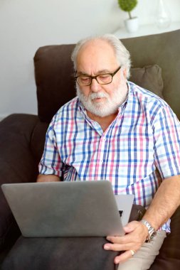 evde kanepede oturan ve dizüstü bilgisayar kullanarak beyaz sakallı üst düzey adam portresi