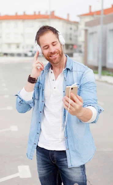 英俊的年轻男子 身穿牛仔衣服听音乐通过耳机和智能手机在街上 — 图库照片