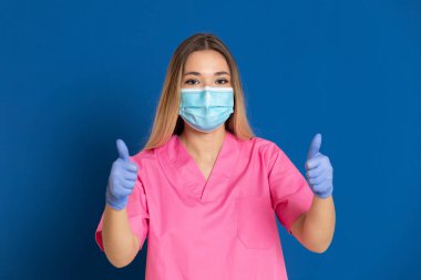 Maske takan genç bir doktor ve mavi arka planda pembe üniformalı.