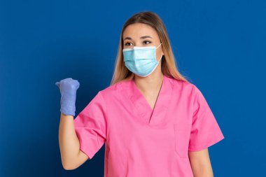 Maske takan genç bir doktor ve mavi arka planda pembe üniformalı.
