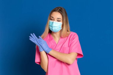 Maske takan genç bir doktor ve mavi arka planda pembe üniformalı.