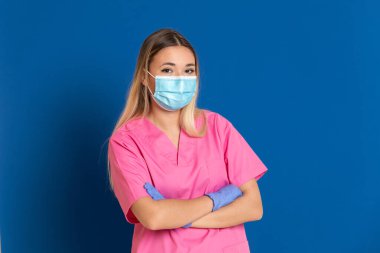 Maske takan genç bir doktor ve mavi arka planda pembe üniformalı.