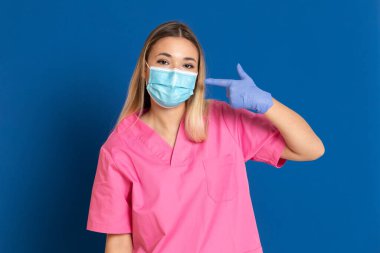 Maske takan genç bir doktor ve mavi arka planda pembe üniformalı.