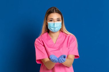 Maske takan genç bir doktor ve mavi arka planda pembe üniformalı.