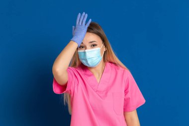 Maske takan genç bir doktor ve mavi arka planda pembe üniformalı.