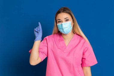 Maske takan genç bir doktor ve mavi arka planda pembe üniformalı.