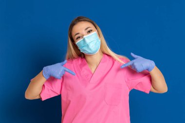 Maske takan genç bir doktor ve mavi arka planda pembe üniformalı.