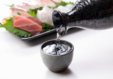 Sake beyaz arkaplana karşı hazırlanmış bir sake bardağına dökülüyor. Sashimi arkada..