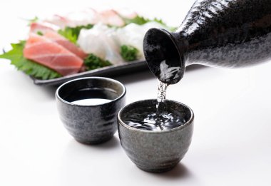 Sake beyaz arkaplana karşı hazırlanmış bir sake bardağına dökülüyor. Sashimi arkada..