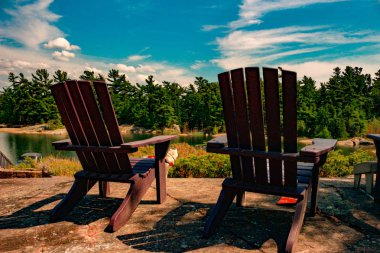 Sakin bir göle bakan bir ahşap iskelede oturup iki Muskoka sandalye. Yeşil ağaçlar arasında yer alan bir beyaz yazlık sudur arasında.