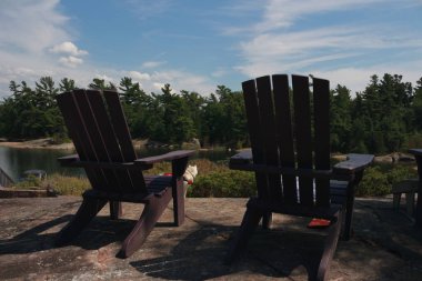Bir göle bakan bir ahşap iskelede oturup iki Muskoka sandalye. Sakin arasında yeşil ağaçlar arasında yer alan bir beyaz yazlık sudur. Yazlık önünde su üzerinde bir tekne iskelesi.