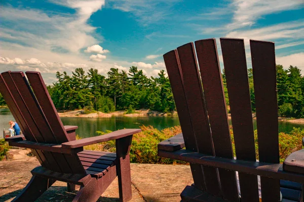 Two Muskoka chairs sitting on a wood dock facing a lake. Across