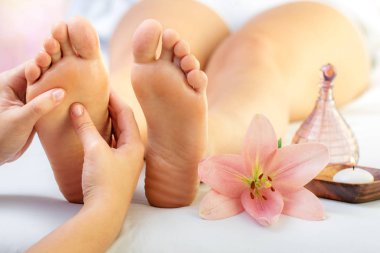 Close up of hands doing foot reflexology massage on female foot. Flower, candle and massage oil next to feet. clipart