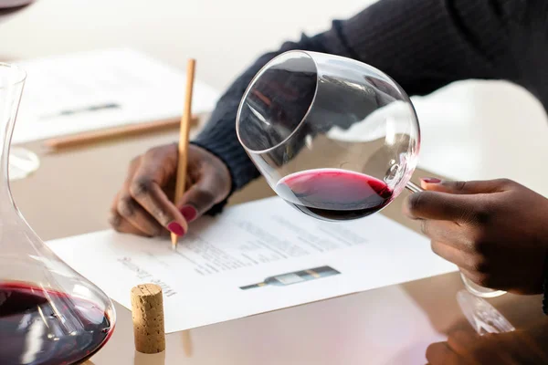 赤ワインのガラスを保持しているアフリカの手の詳細を閉じます ワイン愛好家は ワインの試飲で赤ワインを評価します 女性テーブルでデキャンタでメモを取る — ストック写真