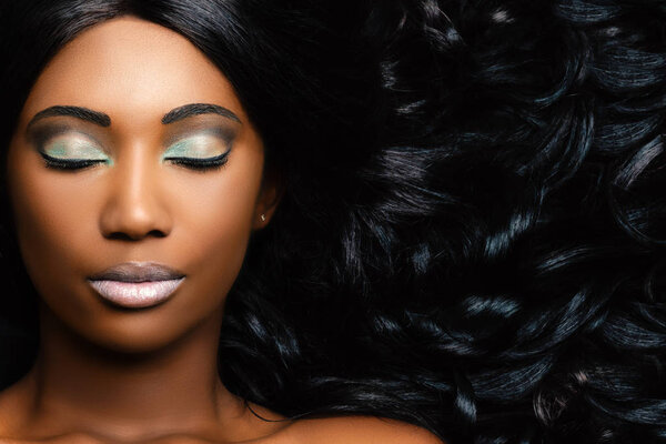 Extreme close up beauty portrait of beautiful young african woman with professional make up. Girl with eyes closed and long curly hair next to face.