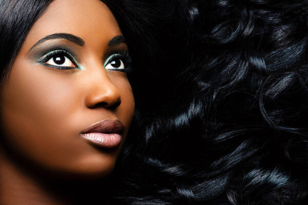 Extreme close up beauty portrait of beautiful young african woman with professional make up. Girl looking at corner with long curly hair next to face.