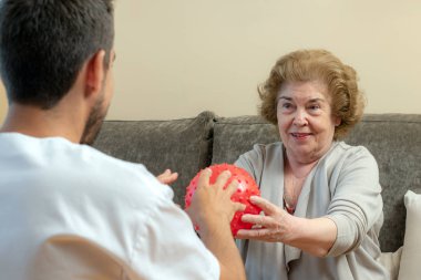 Close up portrait of senior woman doing coordination exercise with male physiotherapist. Old lady catching red plastic ball clipart