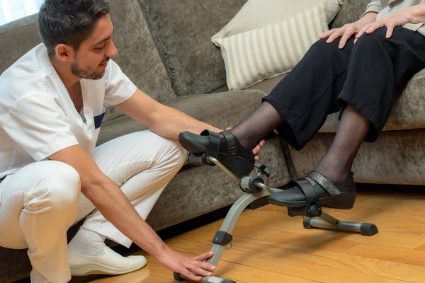 Close up portrait of young male nurse assisting senior patient with rehabilitation pedal.Woman sitting on couch doing exercise with stationary bicycle.