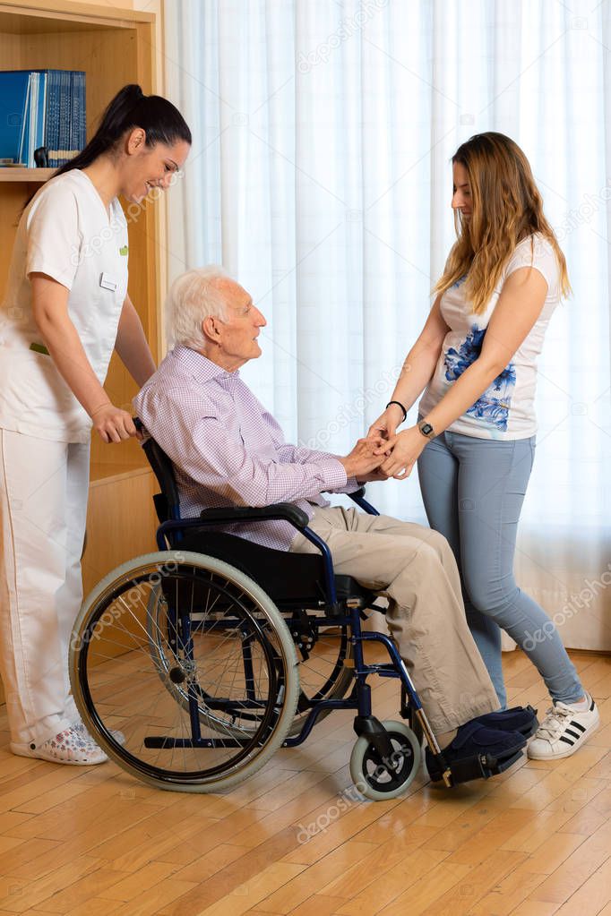 Full length portrait of senior disabled man in wheelchair. Granddaughter and nurse assisting and showing affection to old man in geriatric.
