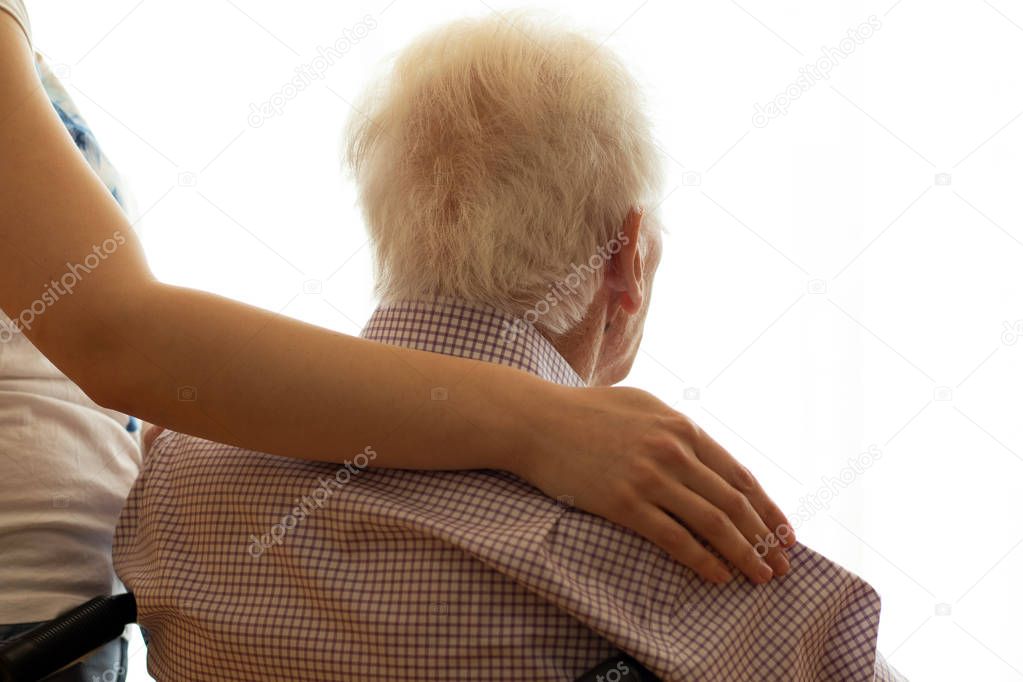 Close up detail of female arm embracing old man. Rear view of senior with window back light. 
