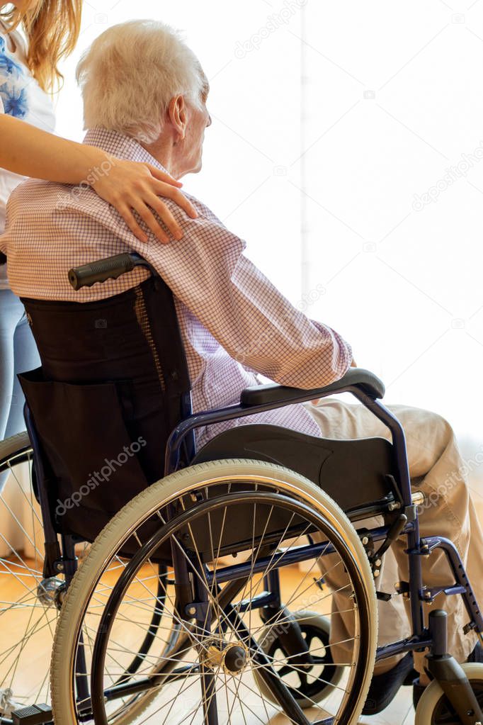 Close up vertical shot of old man in wheelchair staring at window. Female caretaker standing with hand on his shoulder.